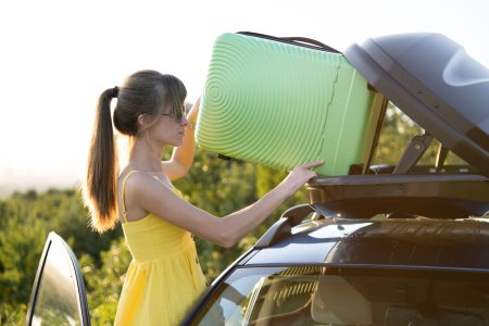 Young woman driver putting green suitcase inside car roof rack. Travel and vacations concept.